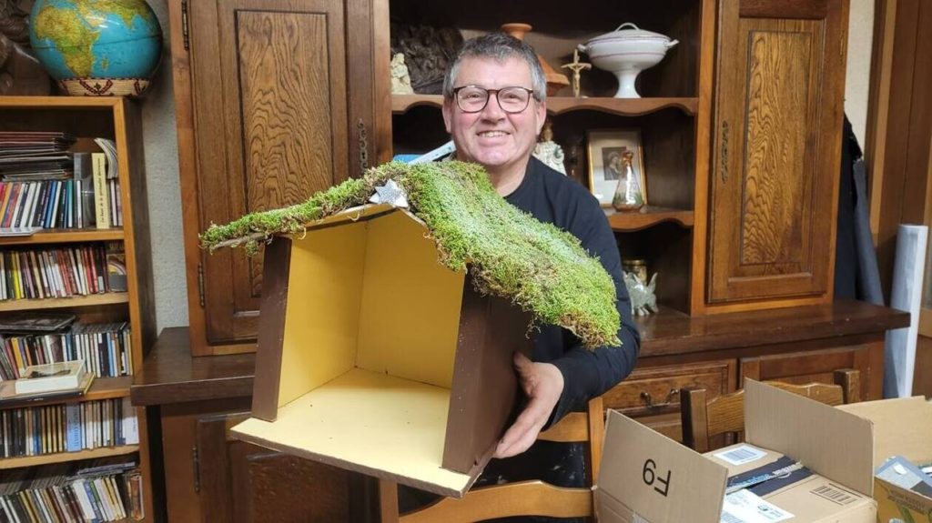Un moine de l'abbaye de la Grainetière souriant, tenant une crèche de Noël en bois faite main, avec un toit recouvert de mousse naturelle. La crèche est posée sur une table, entourée d'autres objets et d'une bibliothèque en arrière-plan.
