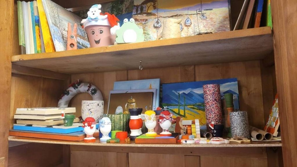 Une étagère en bois contenant divers objets artisanaux réalisés par les moines de l'abbaye de la Grainetière. On y voit des peintures de paysages, des décorations festives comme un pot de fleurs décoré en bonhomme de neige, des coquetiers personnalisés, des objets en bois et en carton, ainsi que des couronnes de Noël.